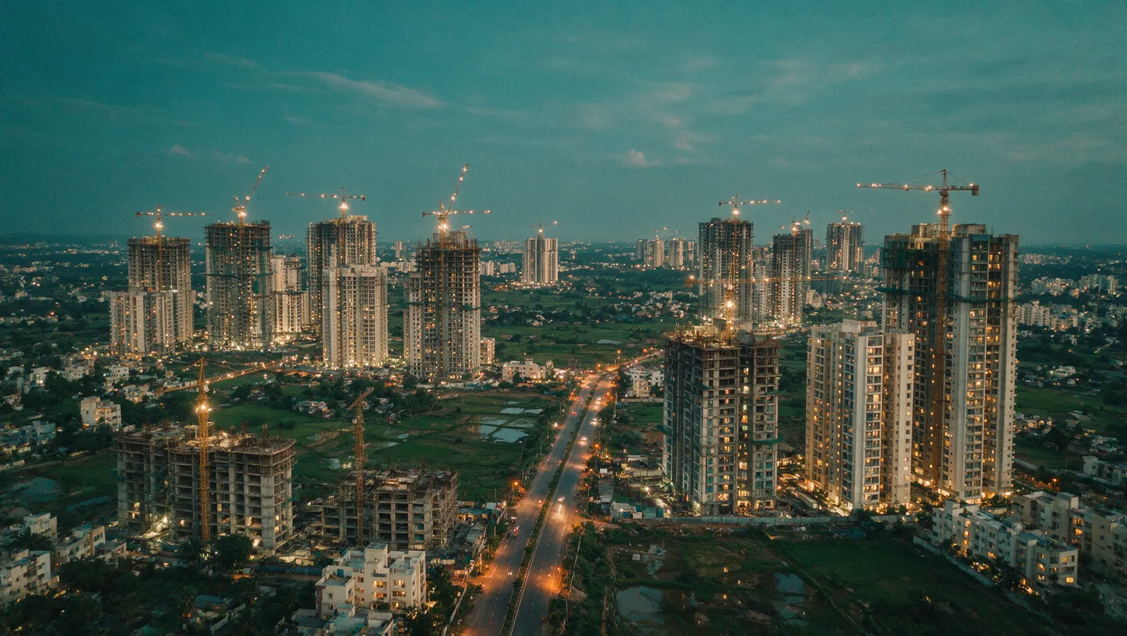 Illustrative editorial image: residential towers in various stages of construction with lit tower cranes across North Bengaluru at early evening